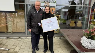 Ein Mann und eine Frau stehen draußen und lächeln. Sie halten ein Zertifikat mit goldenem Rand. Der Mann trägt einen schwarzen Mantel mit Logo. Die Frau hat lange Haare und hält das Zertifikat mit beiden Händen. Hinter ihnen spiegelt eine Glaswand ein Haus und Bäume.