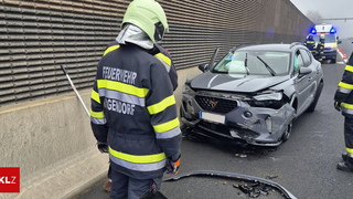 Ein Feuerwehrmann in Uniform steht neben einem beschädigten grauen Auto auf einer Straße. Die Front des Autos ist stark beschädigt, und Trümmer sind auf der Straße verstreut. Der Feuerwehrmann hält eine Kehrschaufel und steht in der Nähe einer Wand.