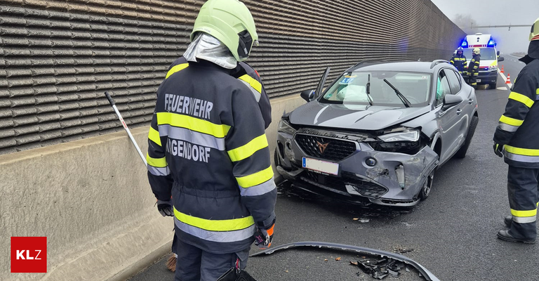 Ein Feuerwehrmann in Uniform steht neben einem beschädigten grauen Auto auf einer Straße. Die Front des Autos ist stark beschädigt, und Trümmer sind auf der Straße verstreut. Der Feuerwehrmann hält eine Kehrschaufel und steht in der Nähe einer Wand.