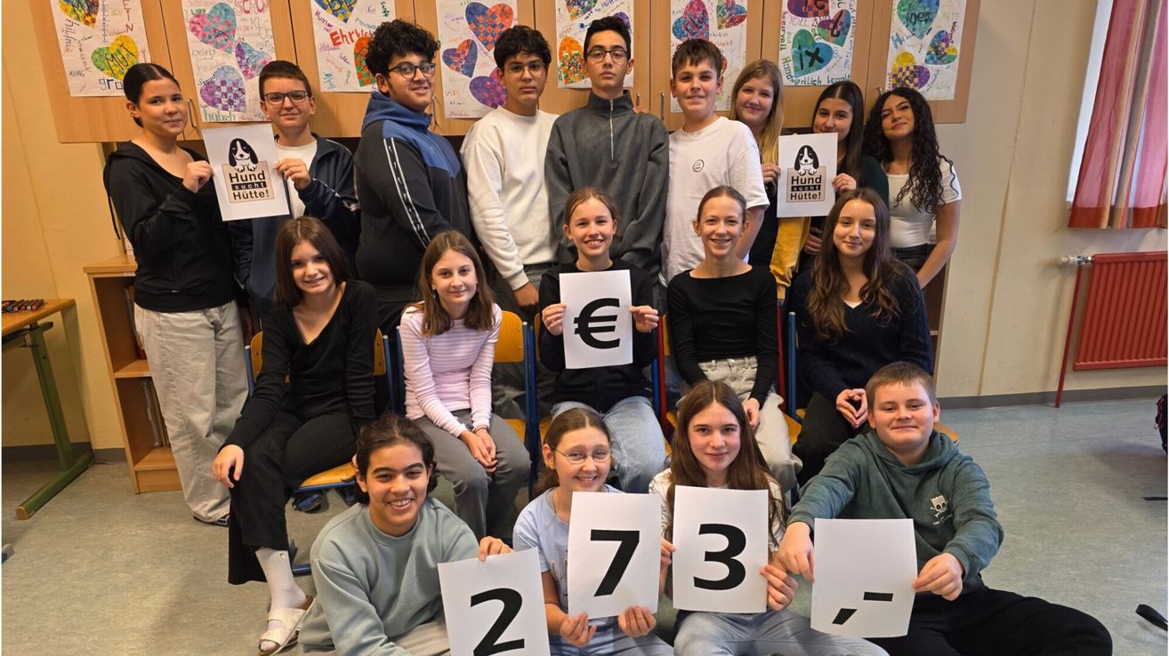 Eine Gruppe von Schülern, die Schilder mit den Zahlen 2, 7, 3 und einem Euro-Symbol halten und für ein Foto in einem Klassenzimmer lächeln.