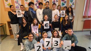 Eine Gruppe von Schülern, die Schilder mit den Zahlen 2, 7, 3 und einem Euro-Symbol halten und für ein Foto in einem Klassenzimmer lächeln.