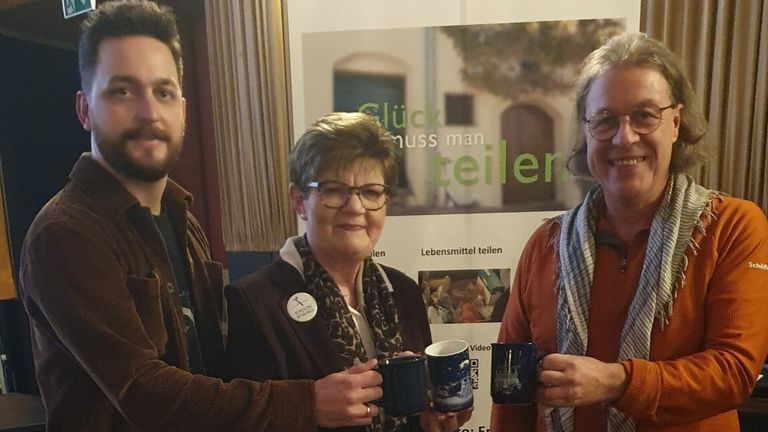 Drei Menschen stehen zusammen und lächeln, während sie Tassen halten. Eine Frau in der Mitte hat ein Abzeichen. Hinter ihnen ist ein Poster mit Text und Bild. Der Hintergrund ist unscharf.