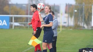Eine Fußballspielerin hält einen Ball und steht auf einem Feld. Hinter ihr hält ein Mann eine Flagge und ein anderer steht. Sie sind alle in Sportbekleidung gekleidet.
