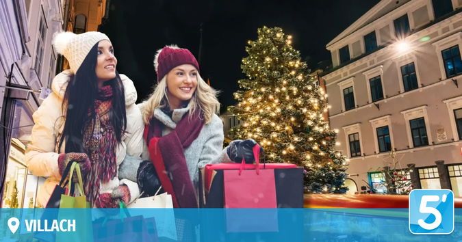 Zwei Frauen in Winterkleidung stehen vor einem Weihnachtsbaum mit Einkaufstüten. Ein Gebäude mit vielen Fenstern befindet sich dahinter. Das Bild hat einen blauen Rand und einen Wasserzeichen mit dem Wort 5'.