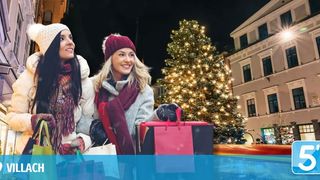 Zwei Frauen in Winterkleidung stehen vor einem Weihnachtsbaum mit Einkaufstüten. Ein Gebäude mit vielen Fenstern befindet sich dahinter. Das Bild hat einen blauen Rand und einen Wasserzeichen mit dem Wort 5'.