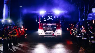 Ein Feuerwehrauto fährt mit eingeschalteten Lichtern durch eine nächtliche Parade. Feuerwehrleute stehen auf beiden Seiten und halten brennende Fackeln.