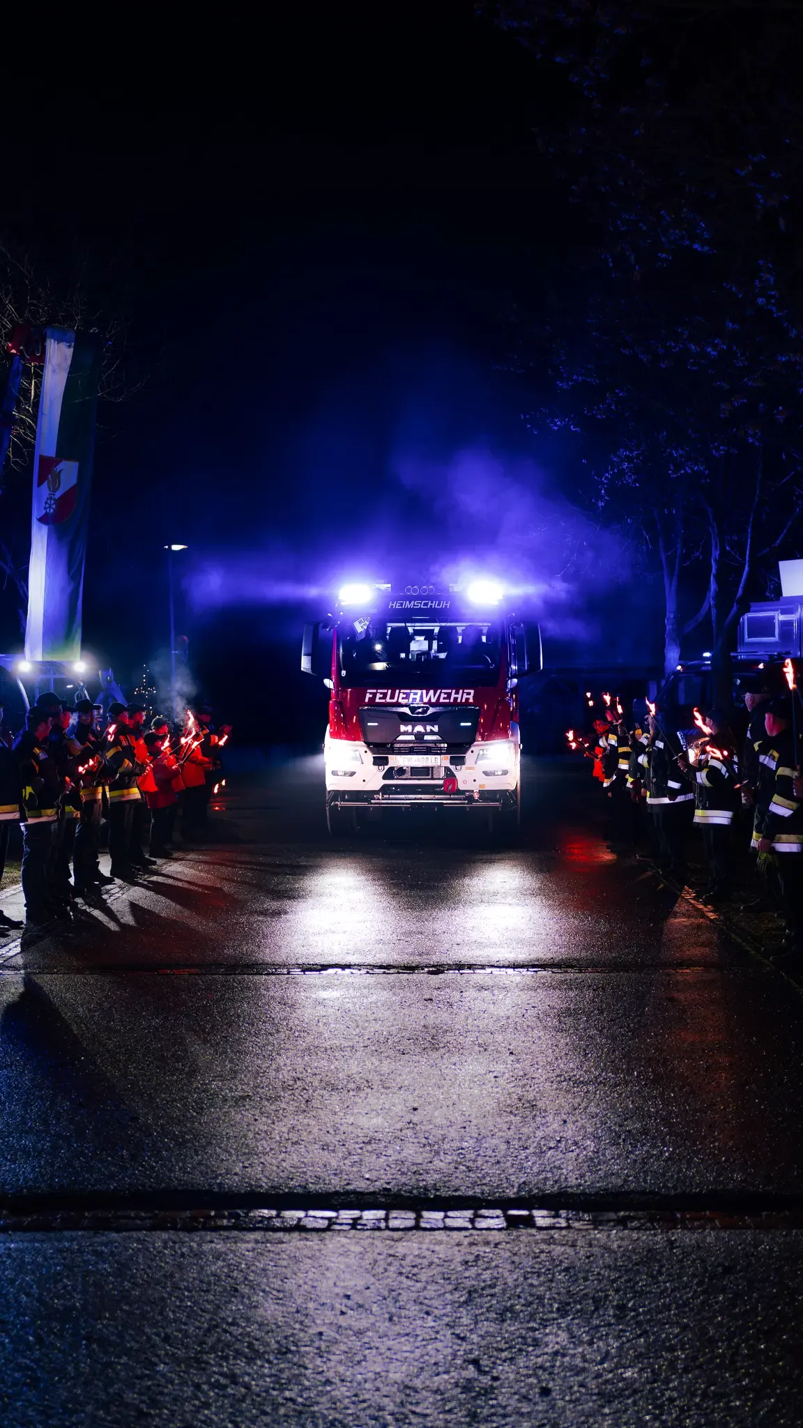 Ein Feuerwehrauto fährt mit eingeschalteten Lichtern durch eine nächtliche Parade. Feuerwehrleute stehen auf beiden Seiten und halten brennende Fackeln.
