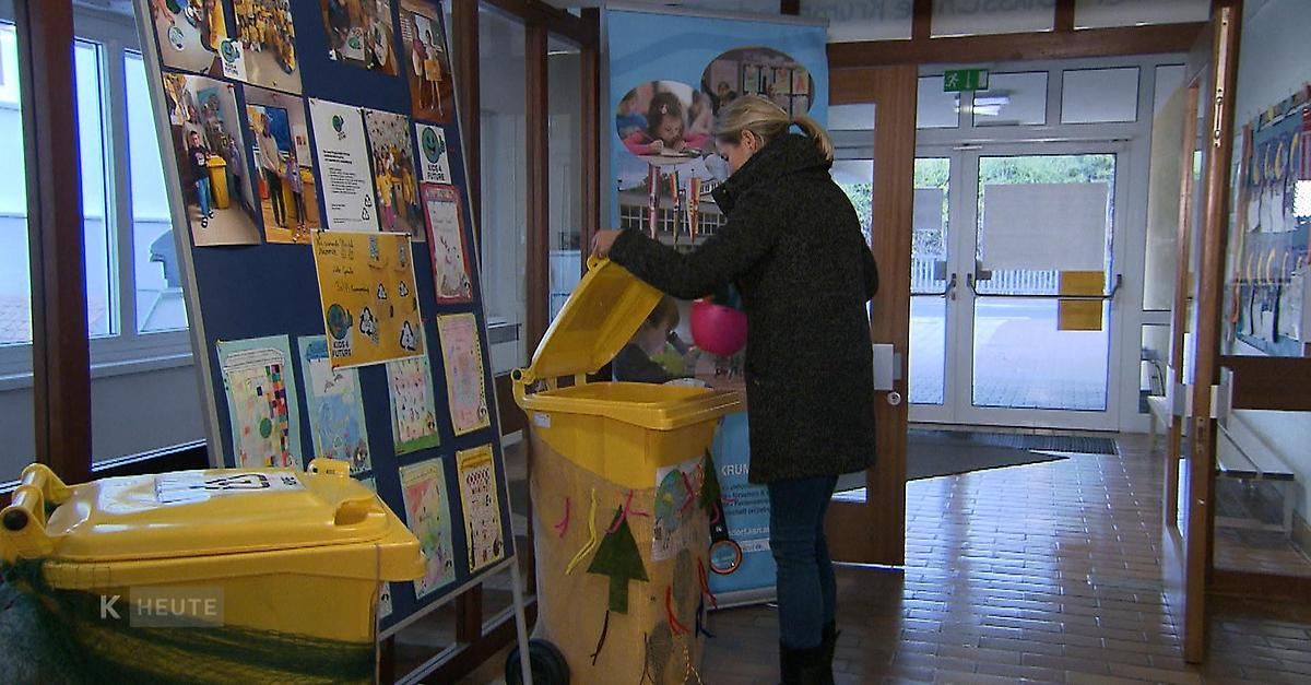 Eine Frau steht vor einem gelben Recyclingbehälter und hält eine rosa Schüssel, möglicherweise wirft sie Abfall hinein. Der Behälter befindet sich in einem Raum mit blauer Wand und Plakaten.