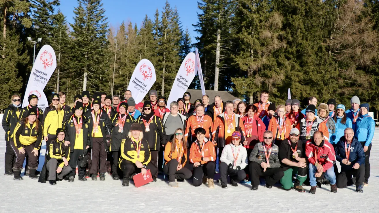 Eine Gruppe von Menschen mit Medaillen und Jacken, die in einem verschneiten Bereich für ein Foto posieren, mit Bäumen und einem Gebäude im Hintergrund.