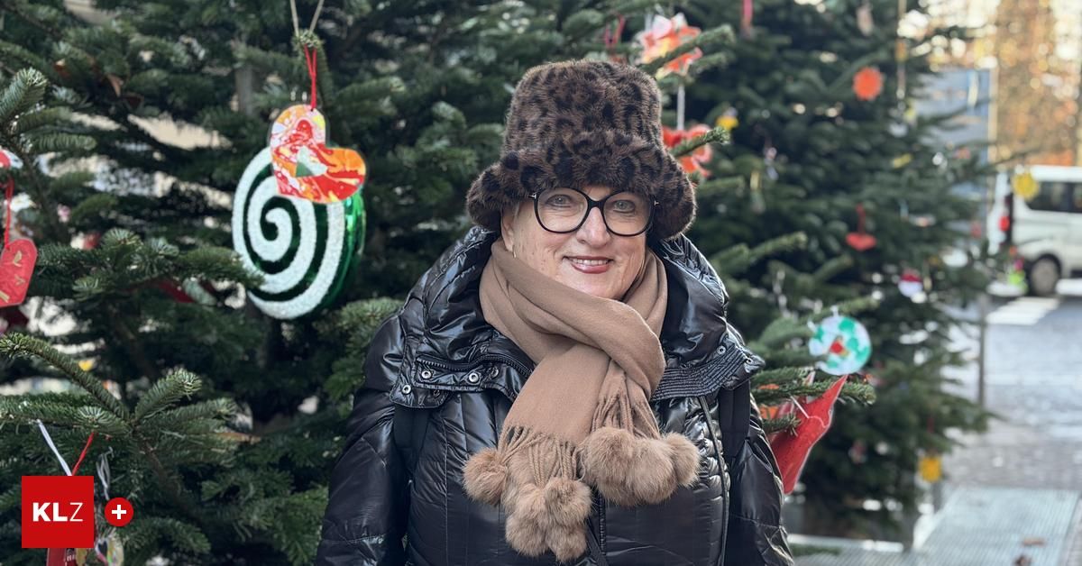 Eine Frau in einem Pelzhut und Schal steht lächelnd vor einem geschmückten Weihnachtsbaum. Der Baum hat Ornamente und der Hintergrund ist leicht unscharf.