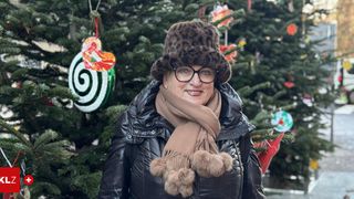 Eine Frau in einem Pelzhut und Schal steht lächelnd vor einem geschmückten Weihnachtsbaum. Der Baum hat Ornamente und der Hintergrund ist leicht unscharf.