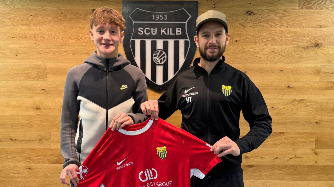 Zwei Männer posieren mit einem roten SCU Kilb-Trikot vor einem Logo an einer Holzwand. Ein Mann trägt eine Kappe und eine schwarze Jacke mit gelbem Logo, der andere ein graues Hoodie.