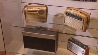 Vier Vintage-Radios sind auf einem Glasregal ausgestellt. Oben links hat ein großes Drehrad, oben rechts ist kleiner, unten links hat ein Philips-Etikett, und unten rechts ist ein kleineres Modell.