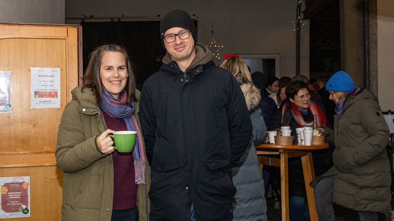Ein Mann und eine Frau stehen lächelnd eng beieinander, drinnen. Die Frau hält eine grüne Tasse. Hinter ihnen sitzen Leute an einem Tisch mit Tassen. Der Hintergrund ist schwach beleuchtet mit Weihnachtsdekoration.