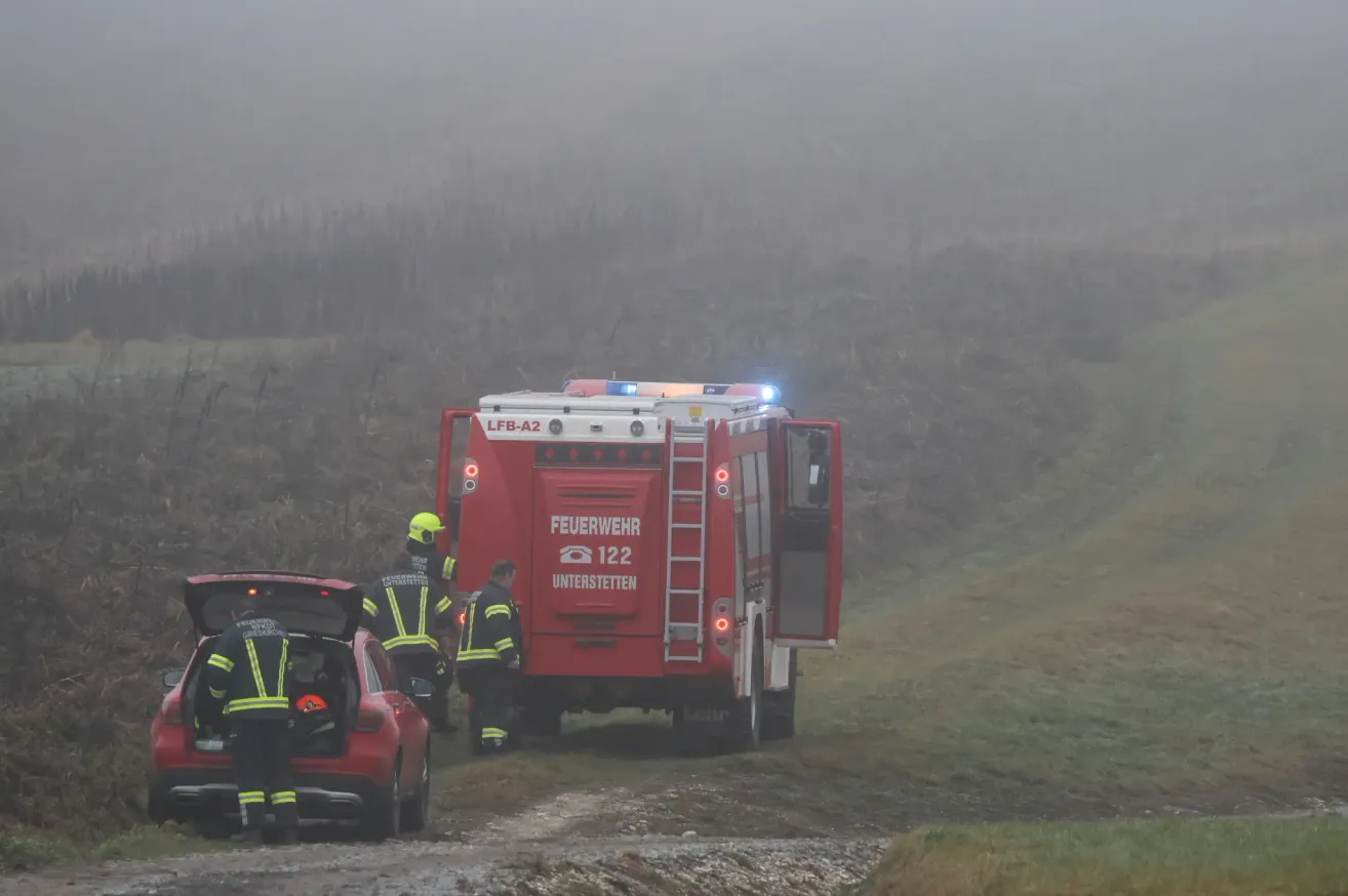 Ein roter Feuerwehrwagen mit der Aufschrift 'Feuerwehr' und der Nummer '122' steht auf einer verregneten Straße. Eine Person mit gelbem Helm steht neben dem Fahrzeug. Eine Person steht am Heck eines roten Autos, und eine weitere Person steht links.