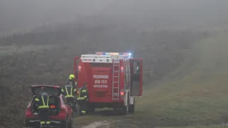 Ein roter Feuerwehrwagen mit der Aufschrift 'Feuerwehr' und der Nummer '122' steht auf einer verregneten Straße. Eine Person mit gelbem Helm steht neben dem Fahrzeug. Eine Person steht am Heck eines roten Autos, und eine weitere Person steht links.