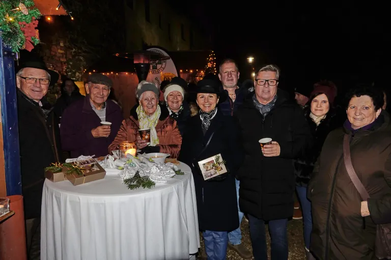 Eine Gruppe älterer Menschen versammelt sich nachts um einen Tisch und posiert für ein Foto. Der Tisch ist mit einer weißen Tischdecke bedeckt und hat verschiedene Gegenstände darauf. Die Menschen tragen Winterkleidung und halten Tassen. Im Hintergrund befinden sich Bäume mit Weihnachtslichtern und ein Banner mit dem Wort 'CORSICA'.