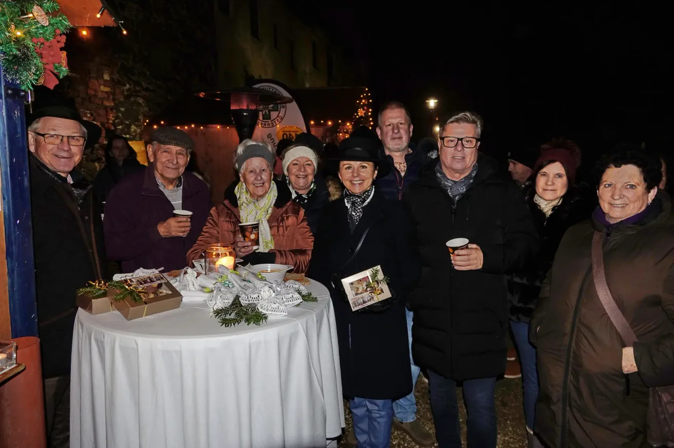 Eine Gruppe älterer Menschen versammelt sich nachts um einen Tisch und posiert für ein Foto. Der Tisch ist mit einer weißen Tischdecke bedeckt und hat verschiedene Gegenstände darauf. Die Menschen tragen Winterkleidung und halten Tassen. Im Hintergrund befinden sich Bäume mit Weihnachtslichtern und ein Banner mit dem Wort 'CORSICA'.