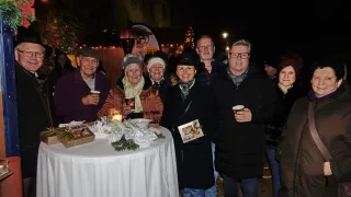 Eine Gruppe älterer Menschen versammelt sich nachts um einen Tisch und posiert für ein Foto. Der Tisch ist mit einer weißen Tischdecke bedeckt und hat verschiedene Gegenstände darauf. Die Menschen tragen Winterkleidung und halten Tassen. Im Hintergrund befinden sich Bäume mit Weihnachtslichtern und ein Banner mit dem Wort 'CORSICA'.