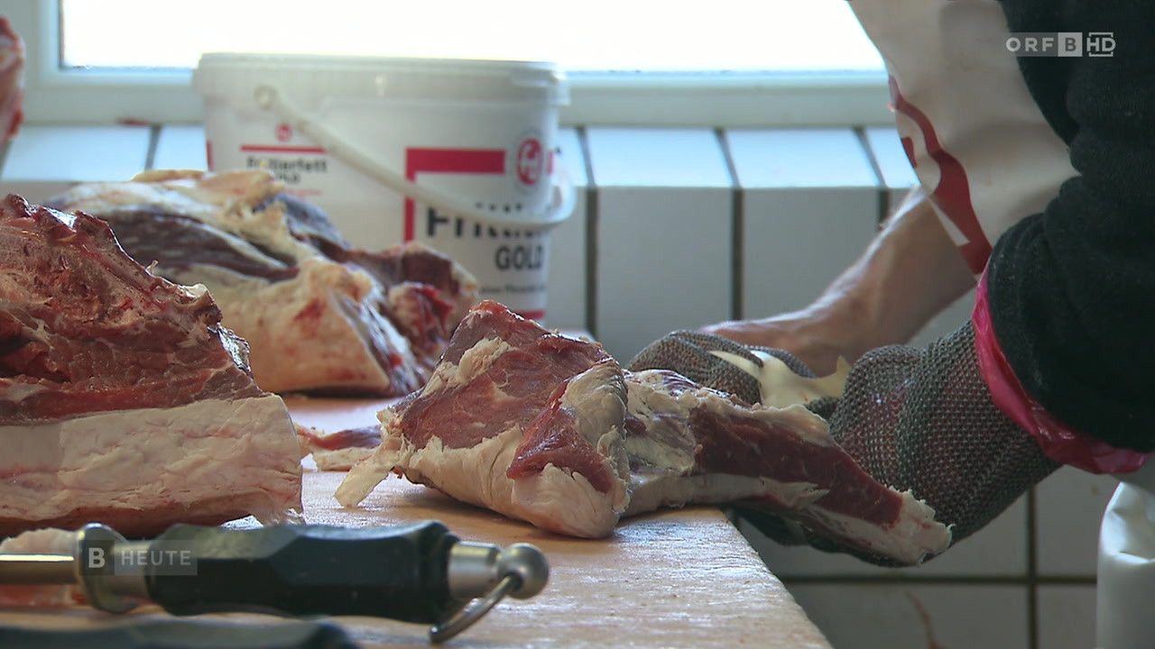 Eine Person, die Handschuhe trägt, schneidet Fleisch auf einem Holzschneidebrett. Ein weißer Eimer mit rotem und schwarzem Text steht hinter dem Fleisch. Ein Werkzeug mit schwarzem Griff und silberner Spitze befindet sich neben dem Fleisch. Die Hände der Person sind verschwommen.