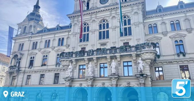 Ein großes weißes Gebäude mit vielen Fenstern und Statuen davor. Eine Uhr ist auf dem Turm sichtbar.