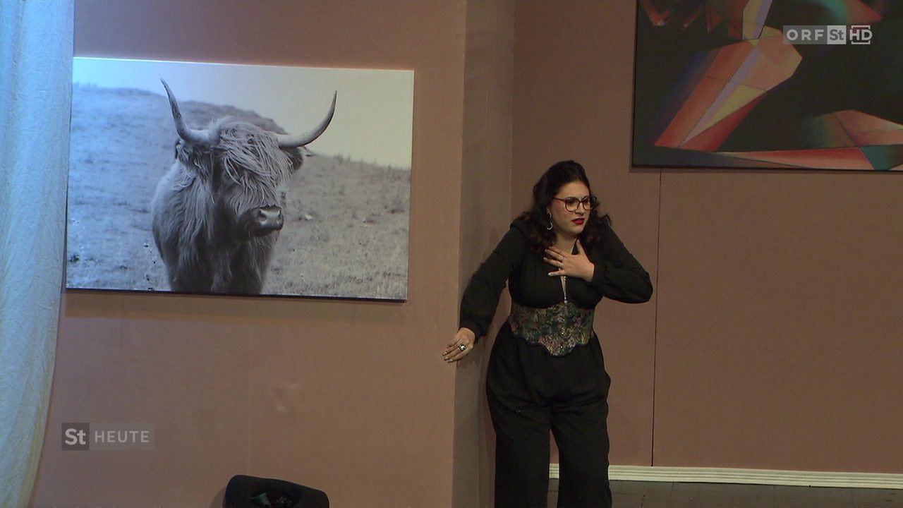 Eine Frau steht in einem Raum mit einem an der Wand montierten Foto eines Hochlandrindes und einem weiteren abstrakten Gemälde, sie trägt eine Brille und einen Ring.