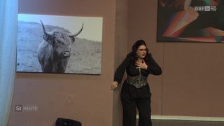 Eine Frau steht in einem Raum mit einem an der Wand montierten Foto eines Hochlandrindes und einem weiteren abstrakten Gemälde, sie trägt eine Brille und einen Ring.