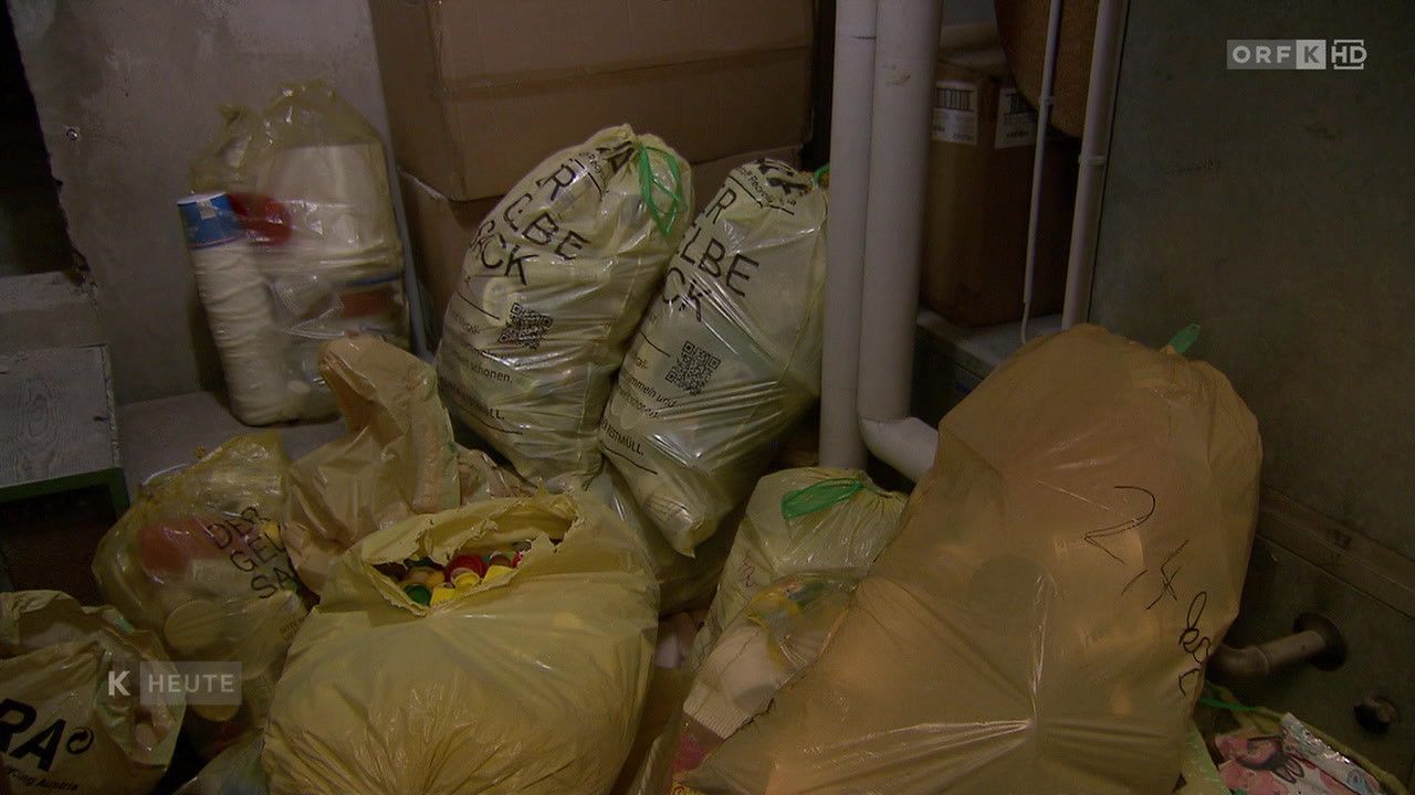 Mehrere gelbe Plastiktüten sind mit kleinen bunten Objekten gefüllt und mit 'Recycling' beschriftet. Dahinter befinden sich Lagerboxen und eine weiße Rohrleitung.