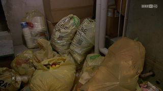 Mehrere gelbe Plastiktüten sind mit kleinen bunten Objekten gefüllt und mit 'Recycling' beschriftet. Dahinter befinden sich Lagerboxen und eine weiße Rohrleitung.