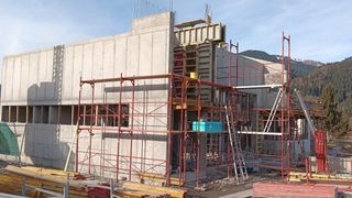 Eine Baustelle mit einer teilweise fertiggestellten Betonwand, roten Gerüsten und einer Leiter. Materialien und Werkzeuge sind herumgestreut.