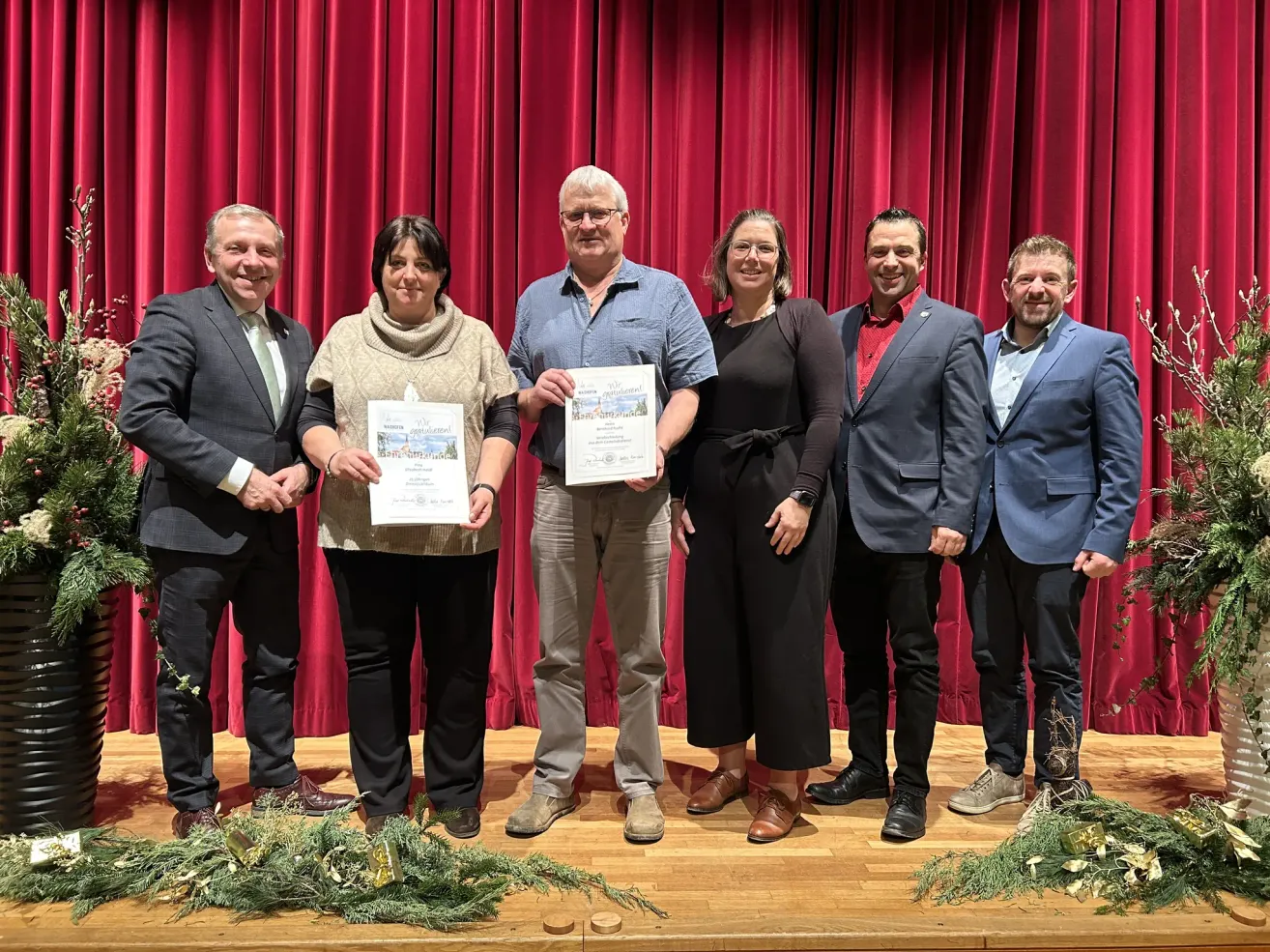 Eine Gruppe von sechs Personen steht auf einer Bühne mit roten Vorhängen im Hintergrund und hält Urkunden in den Händen. Sie sind in formeller Kleidung gekleidet, der Mann links trägt einen Anzug mit Krawatte.