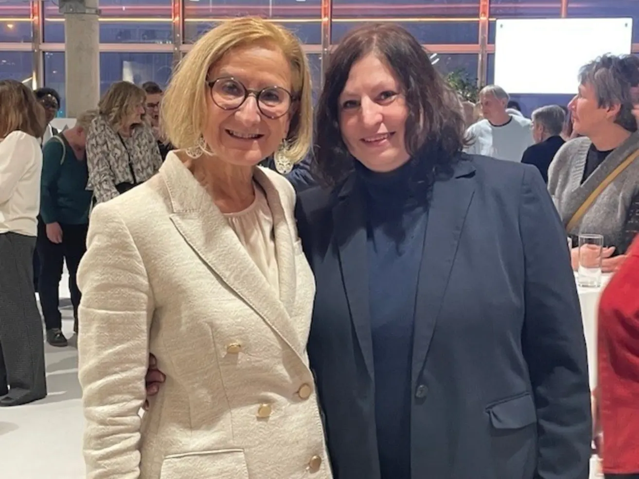 Zwei Frauen stehen lächelnd in einem Raum, eine in einem beigen Blazer und die andere in einem dunkelblauen Anzug. Sie scheinen sich bei einer formellen Veranstaltung zu befinden, mit mehreren Personen hinter ihnen.