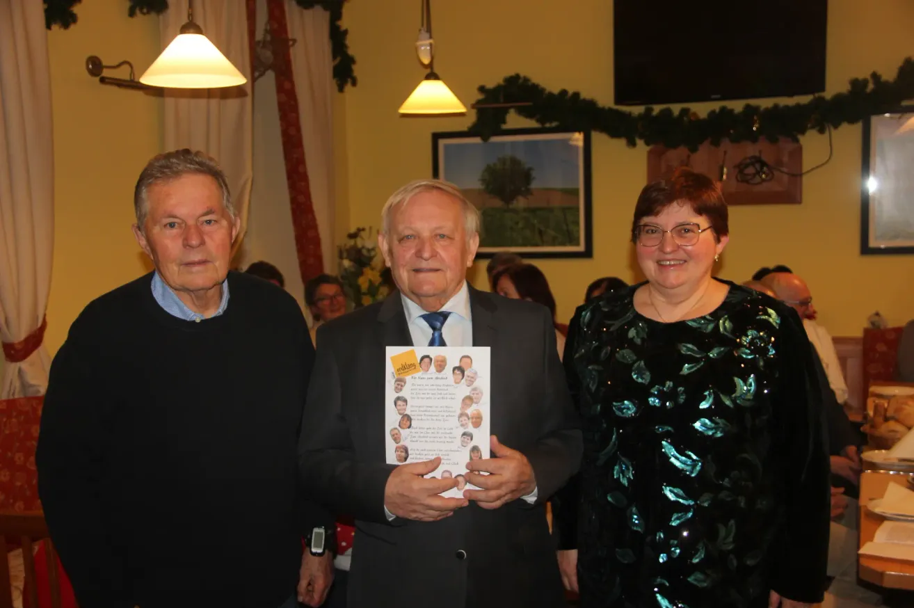 Drei Menschen stehen zusammen; ein Mann hält ein Papier mit Fotos und Text in der Hand und lächelt. Dahinter eine gelbe Wand, ein Bilderrahmen und eine Girlande.