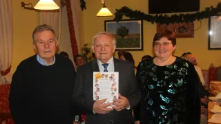 Drei Menschen stehen zusammen; ein Mann hält ein Papier mit Fotos und Text in der Hand und lächelt. Dahinter eine gelbe Wand, ein Bilderrahmen und eine Girlande.