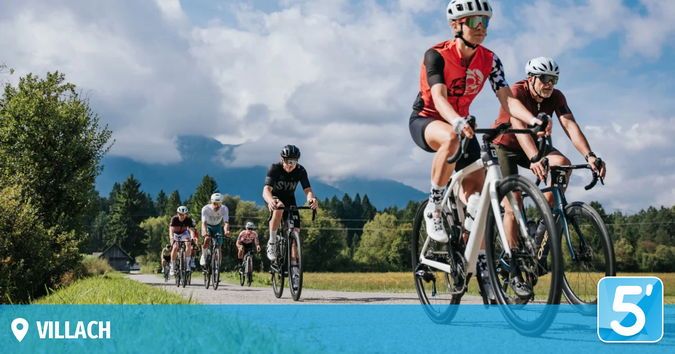 Eine Gruppe von Radfahrern fährt auf einer Landstraße mit Bergen im Hintergrund. Sie tragen Helme und Radtrikots.