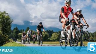 Eine Gruppe von Radfahrern fährt auf einer Landstraße mit Bergen im Hintergrund. Sie tragen Helme und Radtrikots.