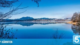 Ein ruhiger See spiegelt den blauen Himmel und die verschneiten Berge wider. Bäume und Gebäude befinden sich auf der anderen Seite des Sees.