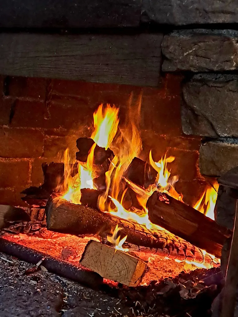 Ein Backsteinkamin mit einem brennenden Feuer, umgeben von Holzscheiten, Steinen und einem hölzernen Kaminsims, strahlt eine warme Glut aus.