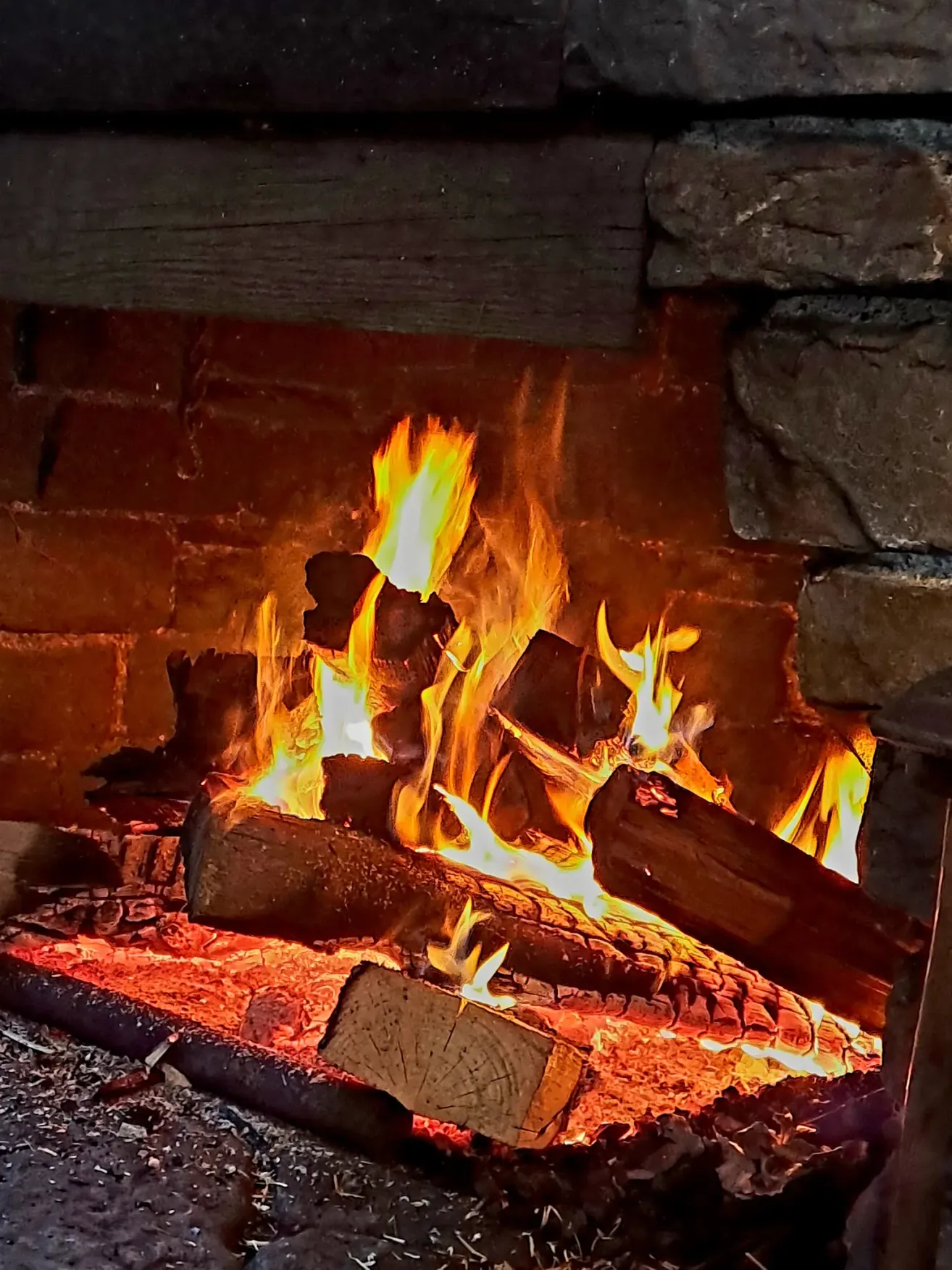 Ein Backsteinkamin mit einem brennenden Feuer, umgeben von Holzscheiten, Steinen und einem hölzernen Kaminsims, strahlt eine warme Glut aus.