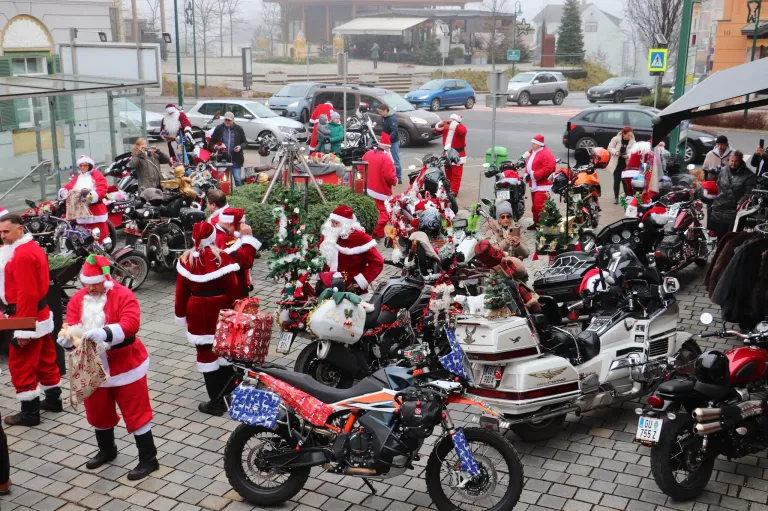 Eine Gruppe von Menschen, die als Weihnachtsmann gekleidet sind, versammelt sich um Motorräder, einige mit Weihnachtsdekoration. Sie stehen auf einer gepflasterten Straße, mit geparkten Autos in der Nähe und einem nebligen Hintergrund.