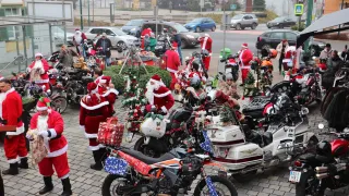 Eine Gruppe von Menschen, die als Weihnachtsmann gekleidet sind, versammelt sich um Motorräder, einige mit Weihnachtsdekoration. Sie stehen auf einer gepflasterten Straße, mit geparkten Autos in der Nähe und einem nebligen Hintergrund.