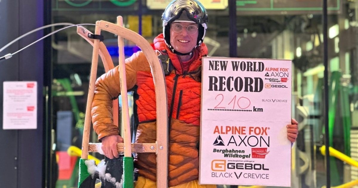 Ein Mann in Skiausrüstung hält ein Schild mit der Aufschrift 'Neuer Weltrekord 210 km' und trägt einen Helm und eine Schutzbrille. Er lächelt und steht vor einem verschneiten Hintergrund.