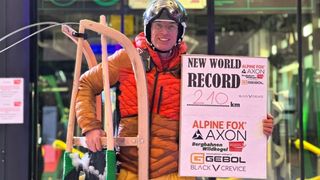 Ein Mann in Skiausrüstung hält ein Schild mit der Aufschrift 'Neuer Weltrekord 210 km' und trägt einen Helm und eine Schutzbrille. Er lächelt und steht vor einem verschneiten Hintergrund.