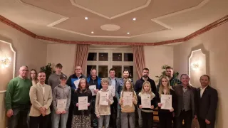 Eine Gruppe von Personen steht zusammen und hält Urkunden in der Hand, mit einem Mann in einem Anzug auf der linken Seite, einer Frau mit Hut in der Mitte und anderen in Freizeitkleidung. Sie befinden sich in einem Raum mit dekorierter Decke, Fenstern und einem Topf.