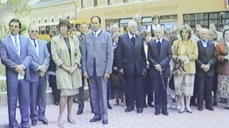 Eine Gruppe formell gekleideter Personen steht vor einem Gebäude namens 'Gebhard-Bauer-Gastzimmer.' Dahinter befinden sich mehrere Menschen, die einen Menschenauflauf bilden.