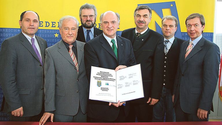 Fünf Männer stehen vor einem gelben Hintergrund. Der Mann in der Mitte hält ein Zertifikat, und sie sind alle in Anzug gekleidet. Der Hintergrund hat ein blaues und gelbes Design und einige Text.