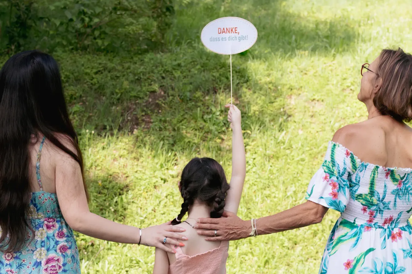 Ein junges Mädchen hält ein Schild mit der Aufschrift 'Danke, dass es dich gibt!' während zwei Erwachsene hinter ihr stehen, einer mit einem Ring und der andere mit einer Armbanduhr.