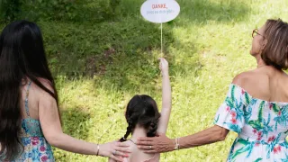Ein junges Mädchen hält ein Schild mit der Aufschrift 'Danke, dass es dich gibt!' während zwei Erwachsene hinter ihr stehen, einer mit einem Ring und der andere mit einer Armbanduhr.