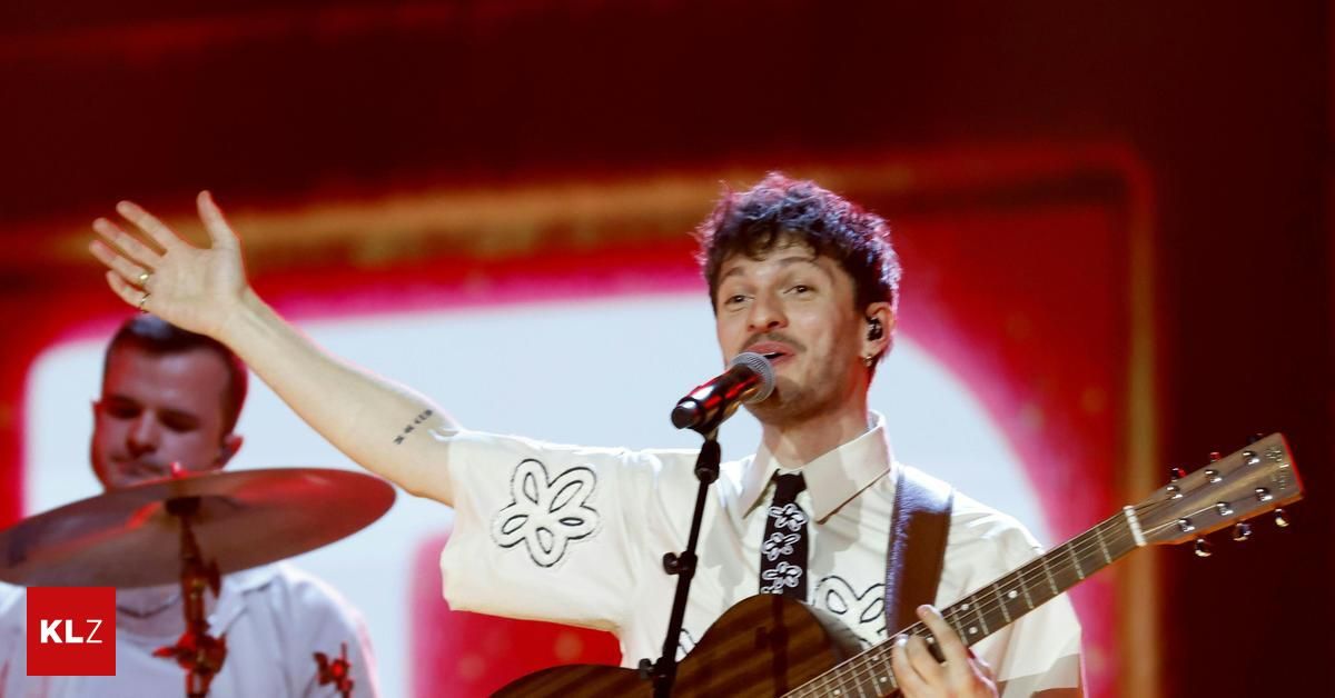 Ein Mann singt in ein Mikrofon und spielt Gitarre. Er trägt ein weißes Hemd mit Blumenmuster und eine schwarze Krawatte. Dahinter spielt ein Schlagzeuger. Die Bühne ist mit roter und weißer Beleuchtung beleuchtet.