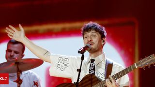 Ein Mann singt in ein Mikrofon und spielt Gitarre. Er trägt ein weißes Hemd mit Blumenmuster und eine schwarze Krawatte. Dahinter spielt ein Schlagzeuger. Die Bühne ist mit roter und weißer Beleuchtung beleuchtet.